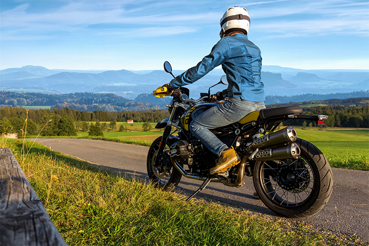 Person wearing a helmet, denim jacket, and yellow shoes sitting on a motorcycle on a country road overlooking rolling hills.
