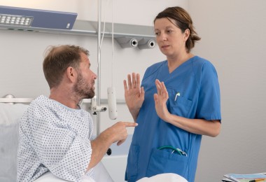 Eine Pflegerin weicht vor einem Patienten zurück, der mit dem Finger auf sie zeigt. Sie hebt abwehrend die Hände. Die Pflegerin trägt einen blauen Kittel. Der Patient ein Krankenhausnachthemd. 
