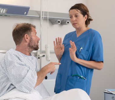 Eine Pflegerin weicht vor einem Patienten zurück, der mit dem Finger auf sie zeigt. Sie hebt abwehrend die Hände. Die Pflegerin trägt einen blauen Kittel. Der Patient ein Krankenhausnachthemd. 