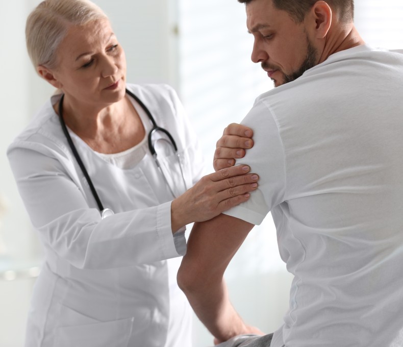 Ärztin in weißem Kittel untersucht die Schulter eines sitzenden Mannes. Sie tastet den Oberarm ab, während der Mann schmerzhaft den Kopf abwendet. Beide tragen weiße Kleidung. Im Hintergrund ist unscharf ein Modell einer Wirbelsäule zu erkennen.