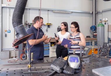In einer Werkstatt mit metallverarbeitender Ausstattung stehen ein Mann und zwei Frauen im Gespräch. Der Mann trägt ein dunkelblaues Poloshirt und erklärt gestikulierend etwas. Die eine Frau hält ein Klemmbrett, die andere ein Werkzeug. Im Vordergrund liegt ein Schweißhelm auf einem großen Metallarbeitstisch, an dem Absaugvorrichtungen und weitere Werkzeuge angebracht sind. Im Hintergrund sind Werkbänke, Gasflaschen und weitere Arbeitsmittel sichtbar.

