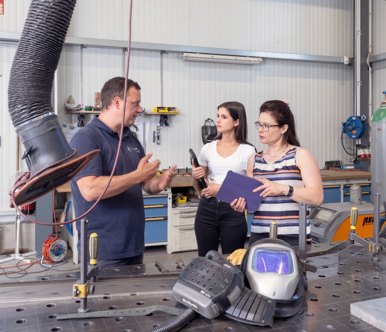 In einer Werkstatt mit metallverarbeitender Ausstattung stehen ein Mann und zwei Frauen im Gespräch. Der Mann trägt ein dunkelblaues Poloshirt und erklärt gestikulierend etwas. Die eine Frau hält ein Klemmbrett, die andere ein Werkzeug. Im Vordergrund liegt ein Schweißhelm auf einem großen Metallarbeitstisch, an dem Absaugvorrichtungen und weitere Werkzeuge angebracht sind. Im Hintergrund sind Werkbänke, Gasflaschen und weitere Arbeitsmittel sichtbar.
