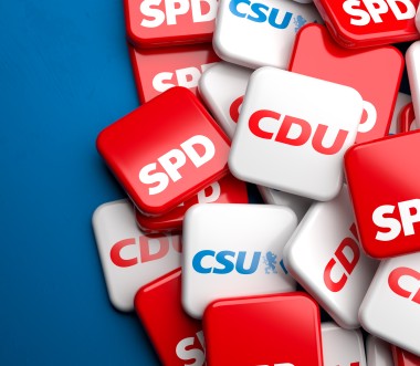 Ein Stapel quadratischer Kacheln mit den Logos deutscher Parteien: SPD (rot), CDU (rot-weiß) und CSU (blau-weiß). Die Kacheln liegen übereinander auf einem blauem Hintergrund, der links ins Bild hineinragt.