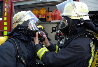 Zwei Feuerwehrleute in vollständiger Schutzausrüstung und Atemschutzmasken stehen vor einem Einsatzfahrzeug. Eine Person hilft der anderen beim Anlegen oder Überprüfen der Atemmaske.