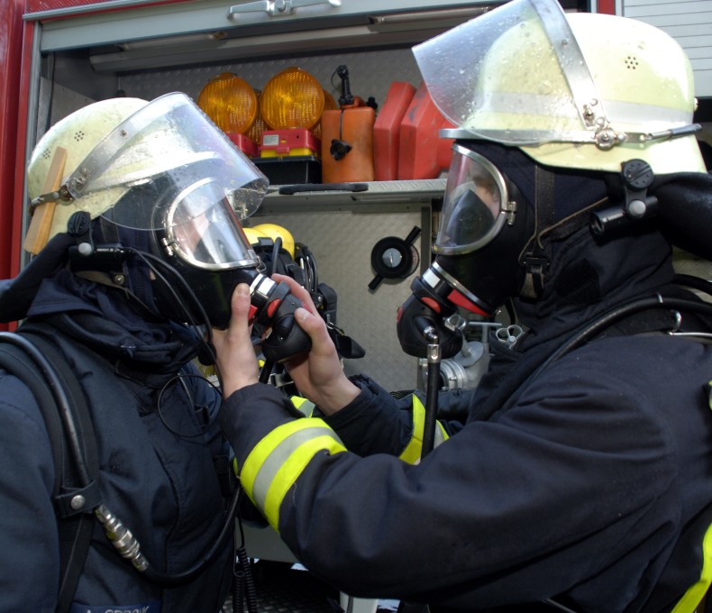Zwei Feuerwehrleute in vollständiger Schutzausrüstung und Atemschutzmasken stehen vor einem Einsatzfahrzeug. Eine Person hilft der anderen beim Anlegen oder Überprüfen der Atemmaske.