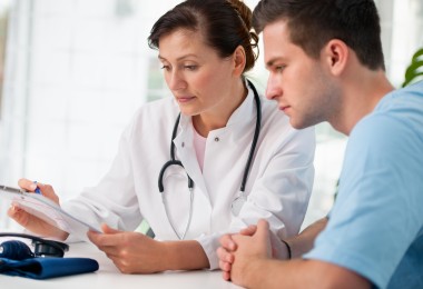 Eine Ärztin in weißem Kittel mit Stethoskop um den Hals sitzt an einem Tisch und bespricht ein Formular mit einem jungen Mann. Beide blicken konzentriert auf das Dokument. Auf dem Tisch liegen ein Blutdruckmessgerät und ein Kugelschreiber.