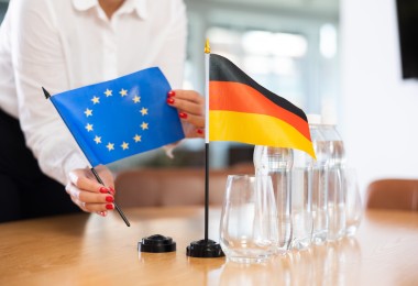 Eine Person im weißen Hemd mit rot lackierten Fingernägeln stellt zwei kleine Tischflaggen auf einem Konferenztisch auf: eine blaue Flagge der Europäischen Union mit gelben Sternen und eine schwarz-rot-goldene Flagge Deutschlands. Daneben stehen mehrere Wasserflaschen und leere Gläser. 
