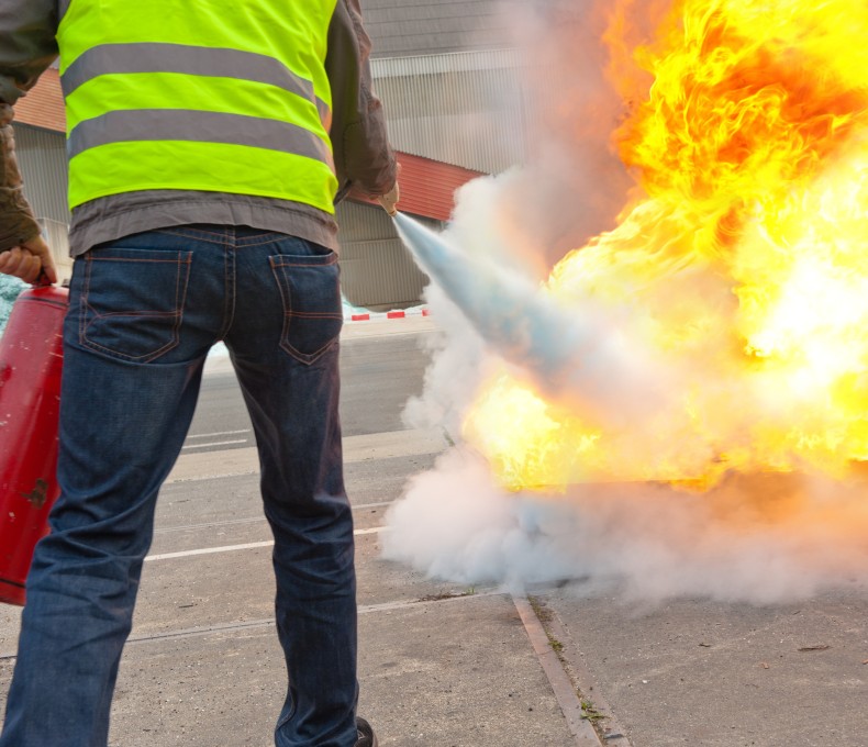 Eine Person mit Warnweste löscht bei einer Brandschutzübung im Freien mit einem Feuerlöscher einen großen, kontrollierten Flammenbrand.
